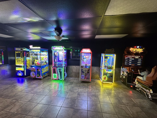 Row of glowing Fun City Games claw machines next to a sit-down racing arcade cabinet inside a partner venue's game room
